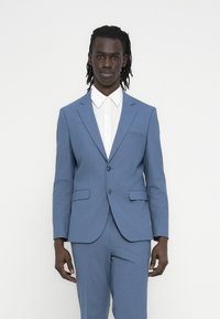 Άνδρας που στέκεται φορώντας ένα μπλε κοστούμι με λευκό πουκάμισο με γιακά, μπροστά σε ένα απλό λευκό φόντο, κοιτάζοντας την κάμερα με ουδέτερη έκφραση.