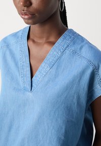 Lyseblå denim top med V-udskæring, korte ærmer og synlige syninger. Glat tekstur med en afslappet pasform.