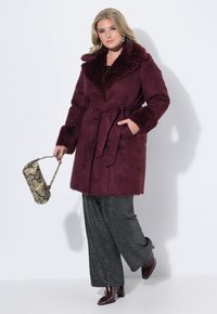 Burgundyröd kappa med pälskrage och manschetter, bälte i midjan och två framfickor. Kombinerad med mörka, glittriga vida byxor och en väska med ormtryck.