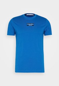 Modré bavlněné tričko s kulatým výstřihem a krátkými rukávy. Na hrudi má černé logo "Calvin Klein Jeans". Hladká textura.