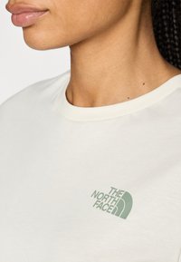 Camiseta de color beige claro con un cuello redondo y una textura suave. La parte delantera presenta un logo verde de "The North Face" en el lado izquierdo.
