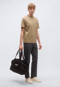 Bézs színű, környakú pamut póló sötétszürke nadrággal kombinálva. A modell egy fekete, "BOSS" logóval ellátott táskát tart. Világos színű sportcipő.