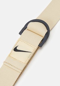 Vaahtokankainen beige vyö mustalla muovisoljella, jossa on pieni suorakulmainen etiketti, jossa on Nike-logo mustalla vaalealla taustalla.