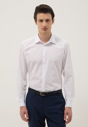 Hombre con cabello oscuro que lleva una camisa blanca metida en pantalones azul marino con cinturón negro, de pie frente a un fondo liso.