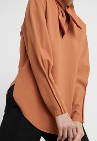 Blouse orange rouille à manches longues avec un détail de nœud au col. Fabriquée en tissu lisse, avec un ourlet incurvé et des fentes latérales.