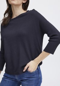 Tumši zila džemperis ar brīvu piegriezumu, plašu apkakli un trīs ceturtdaļu piedurknēm, izgatavots no strukturēta dzijas materiāla. Nēsāts ar zilām džinsām.