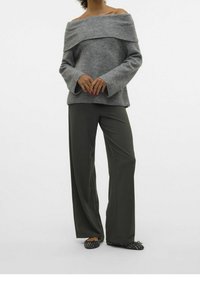 Femme portant un pull gris à épaules dénudées, un pantalon large foncé et des ballerines à motifs, debout les mains jointes.