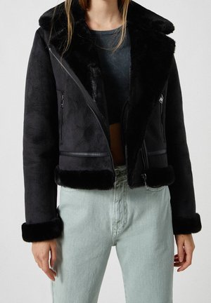 Sort faux fur jakke med bred krave, cropped design, lynlås-lukninger og blød tekstur. Har kontrasterende sort kant og håndledsmanchetter.