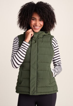 Woman with curly hair smiles, wearing a green quilted sleeveless vest over a black and white striped long-sleeve shirt.