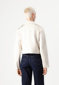 Cream-colored cropped jacket with a collar, long sleeves, and buttoned cuffs, paired with dark blue jeans. Smooth texture with minimal detailing.