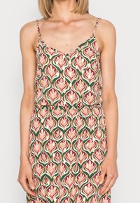 Kvinna i ärmlän klänning med rosa, grön och gyllene blommönster och tunna axelband, stående mot en vit bakgrund.