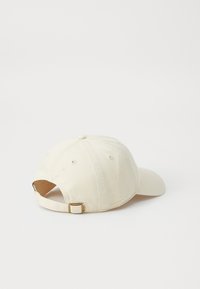 Beige baseball cap made of cotton, featuring a curved brim, stitched eyelets, and an adjustable strap with a metal clasp at the back.