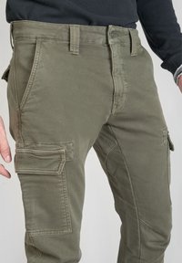 Oliwkowe spodnie cargo z bocznymi kieszeniami noszone z czarną bluzką z długim rękawem, ukazujące talię i górną część nóg.