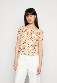 Top floral de manga curta em tecido amarelo suave, com decote volants e padrões de flores vermelhas dispersas, usado com calças brancas.