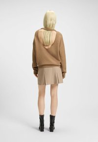 Brauner Strickpullover mit großem offenen Rücken, kombiniert mit einem beigen, plissierten Minirock und schwarzen Blockabsatz-Stiefeletten.