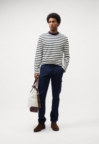 Stribet langærmet skjorte i marineblå og creme, parret med marineblå cargo-bukser. Modellen holder en creme- og brun duffeltaske og har brune ankelstøvler på.