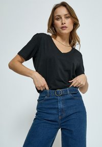 Jonge vrouw draagt een zwart T-shirt met korte mouwen, ingestopt in een hoge taille blauwe jeans met een riem met een ronde gouden gesp.
