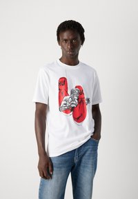 Junger Mann trägt ein weißes T-Shirt mit roten Boxhandschuhen und einem Boxer-Graphic, kombiniert mit blauen Jeans, vor einem schlichten hellen Hintergrund.