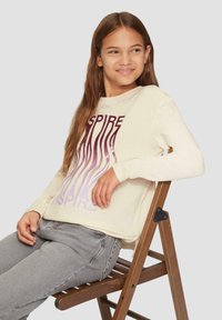 Cremefarbiges Langarmhemd mit einem Farbverlauf-Graphic "INSPIRE" in Burgunderrot und Lavendel. Getragen mit grauen Jeans, sitzend auf einem Holzstuhl.