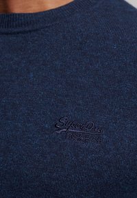 Marineblauer gestrickter Pullover mit Rundhalsausschnitt und gerippten Bündchen, der ein gesticktes Logo in einem dunkleren Farbton auf der linken Brustseite aufweist.