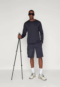 Άνδρας που φοράει γυαλιά ηλίου, μαύρο μπλούζα με μακρύ μανίκι, σορτς και άσπρα αθλητικά παπούτσια, κρατώντας μπαστούνια πεζοπορίας μπροστά σε απλό ανοιχτό φόντο.