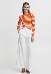 Orange och vitrandig långärmad topp kombinerad med hög midja, vita vida byxor med guldknaffar. Släta texturer.