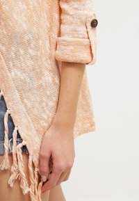 Cardigan tricoté orange clair avec manches retroussées, doté d'un bouton et de franges sur l'ourlet. Fabriqué en matière douce et texturée.