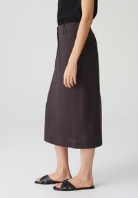 Jupe A-line marron foncé en tissu léger, dotée d'une coupe mi-longue, de poches latérales et d'une fermeture à boutons sur le devant, accompagnée de sandales à enfiler noires.