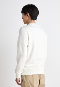 Φούτερ με στρογγυλή λαιμόκοψη, λευκό, από μαλακό ύφασμα, με ριμπ μανσέτες και στρίφωμα, μινιμαλιστική σχεδίαση, πίσω όψη. Φορεμένο με μπεζ παντελόνι.