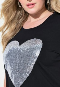 Černé tričko s krátkým rukávem s velkým stříbrným srdcem z flitrů a texturovaným vzorem v oblasti hrudi. Hladká tkanina.