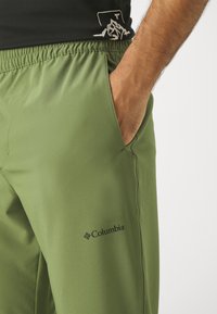 Gröna sportbyxor i lättviktstyg med elastisk midjeresår och sidoficka, med en liten svart Columbia-logotyp.