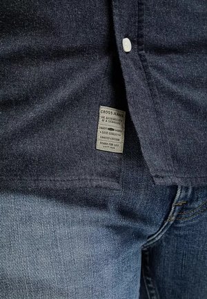 Ciemnoniebieska koszula dżinsowa z zapięciem na guziki, z metką "CROSS JEANS" oraz połączeniem ciemnych i jasnych tekstur denimu.