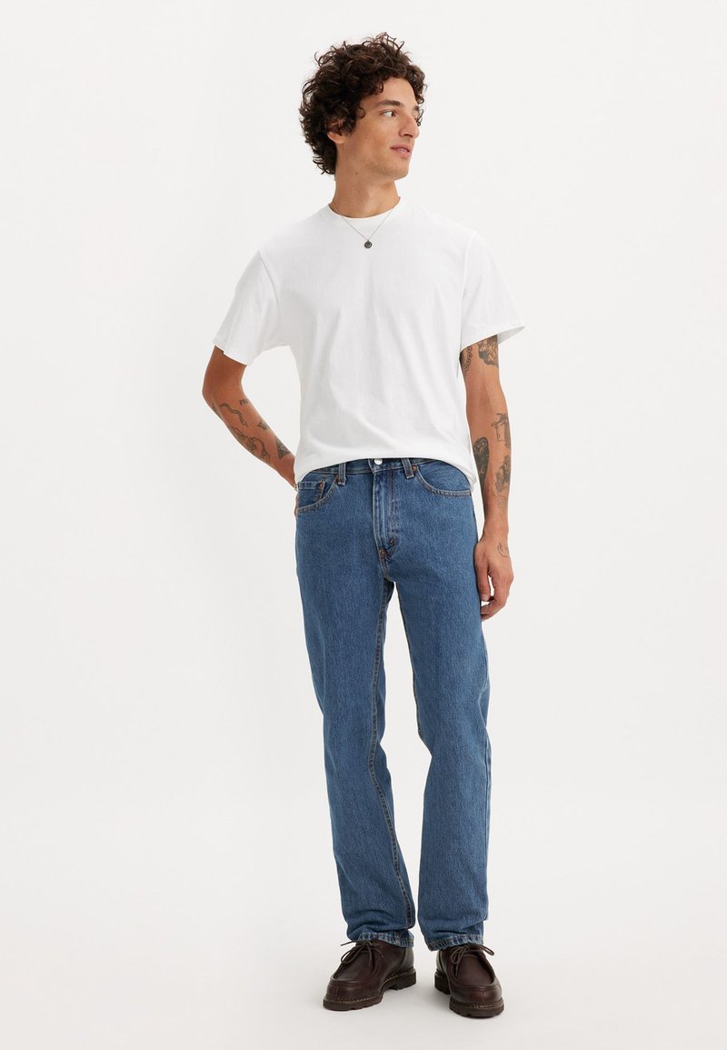 T-shirt blanc à col rond associé à un jean en denim bleu à coupe droite et des chaussures à lacets marron foncé. Des tatouages visibles sur les bras.