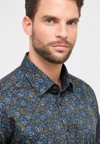 Man met kort bruin haar en een baard, gekleed in een blauw overhemd met bloemenpatroon, kijkend naar de camera.