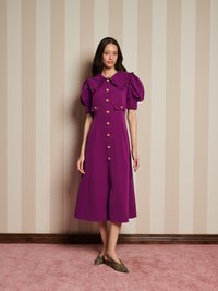 Mulher vestindo um vestido midi roxo com mangas bufantes, botões dourados, colarinho pregueado e sapatos de estampado de leopardo, em pé sobre um tapete rosa ao lado de uma parede riscada.