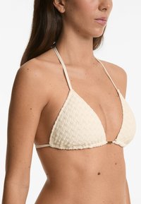 Top bikini di colore crema con un motivo testurizzato a maglia. Presenta spalline sottili regolabili e un piccolo elemento dorato al centro.