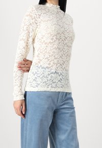 Spetsblus med lång ärm i krämigt off-white med blommönster. Har hög hals och texturerad finish, i kombination med ljusblå kordbyxor.