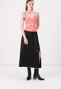 Coral crop top with thin straps and a decorative band, paired with a long black satin skirt featuring a side slit and black ankle boots.