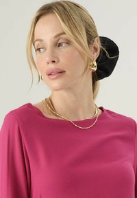 Maglia fucsia a maniche lunghe con scollatura rotonda, collane dorate a strati e un elastico per capelli nero, con capelli lisci e definiti.
