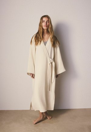 Woman standing barefoot against beige wall wearing long, cream-colored waffle-textured robe with wide sleeves tied at the waist, long hair down.
