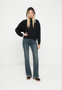 Svart kort hoodie med hette, løs passform; kombinert med medium blå utsvingte jeans. Modellen har på seg svarte sko med lyse såler.