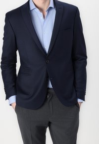 Granatowa, dopasowana marynarka z jednym guzikiem, klapami w notch, i dwiema przednimi kieszeniami, zestawiona z jasnoniebieską koszulą i szarymi spodniami.