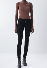Bodysuit marrón de canalé con detalle de un solo hombro, combinado con jeans skinny negros de cintura alta que cuentan con un cierre de botón y bolsillos frontales.