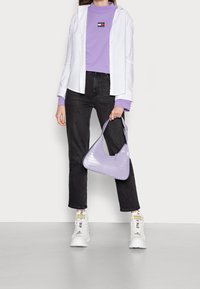 Bolso de mano texturizado en color púrpura con forma curva, combinado con una camisa blanca de botones sobre un top lila y jeans negros, complementado con zapatillas blancas.