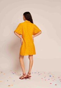 Vestido amarelo de rendas com mangas curtas e evasê e colarinho. O padrão apresenta um design floral. Usado com sapatos de salto vermelho com tiras. Confetes no chão.