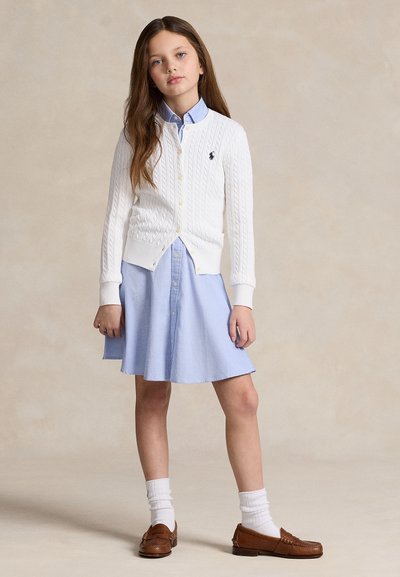 Jeune fille debout, portant un cardigan en maille blanc par-dessus une robe bleue à boutons, des chaussettes blanches et des mocassins marron sur un fond beige uni.
