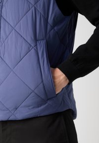 Világoskék steppelt mellény gyémántmintával, oldalzsebbel és alatta egy fekete long-sleeve pólóval. Simább, párnázott textúrájú.