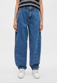 Μπλε τζιν παντελόνια με φαρδιά μπατζάκια, ψηλή μέση, μεγάλες τσέπες και διακριτικό ξεθώριασμα, σε συνδυασμό με λευκά αθλητικά παπούτσια για αντίθεση.