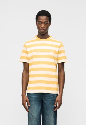 Jonge man die naar voren kijkt, gekleed in een geel-wit gestreept "GUESS JEANS" T-shirt en een blauwe jeans, tegen een effen lichte achtergrond.