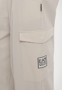 Μπεζ cargo παντελόνια κατασκευασμένα από μαλακό ύφασμα, διαθέτοντας μία μεγάλη πλαϊνή τσέπη και ένα λογότυπο σε ασπρόμαυρο χρώμα στο μπροστινό μέρος.